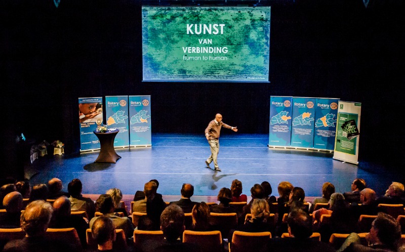 Luc van Bussel op een groot podium met publiek en groot scherm met tekst 'kunst van verbinding' inzake Waarom Hospitality - Hoe kan ik je inspireren?