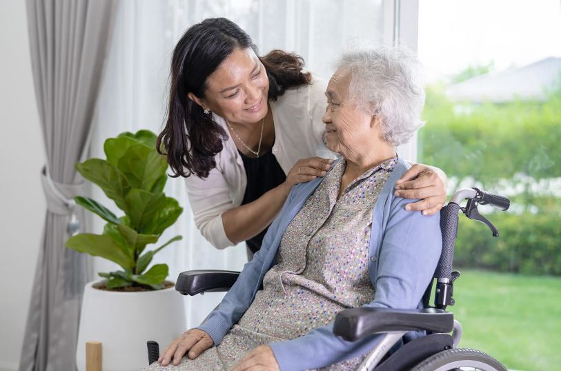 Intrinsieke Gastvrijheid: Glimlachende dame van middelbare leeftijd stelt oudere grijze dame in rolstoel gerust door middel van armen op haar schouders te leggen.