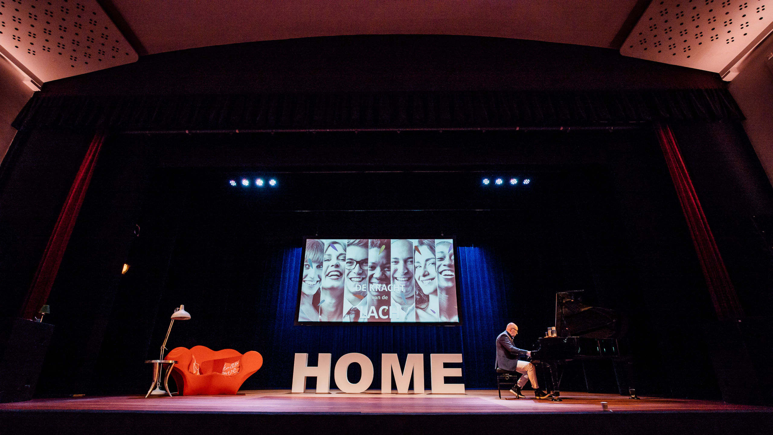 Luc van Bussel op podium achter piano in een theater. Mensen voelen zich als het ware thuis. Gastvrij at Home - inspiratie door Luc