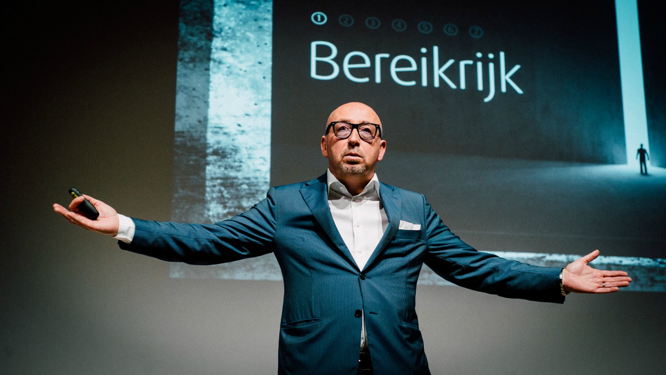 Luc van Bussel - Gastvrijheidsmissie - Luc op podium in groen pak met armen gespreid met op achtergrond presentatie over bereikrijk.